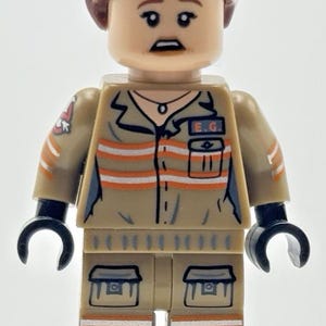 May include: A Lego minifigure with brown hair and a tan jumpsuit featuring orange reflective stripes, a black collar, and a pocket with the letters E.G. printed on the chest. The figure has black hands and dark blue boots with orange trim.