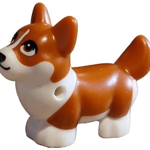 May include: A small plastic toy dog figurine featuring a brown and white coat pattern. The figure has pointed ears, a short tail, and a white muzzle with a black nose and brown eyes. A small circular hole is visible on the side of the neck area.
