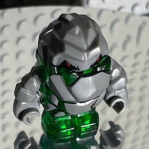 Może przedstawiać: Szara i zielona minifigurka Lego o skalistym, opancerzonym designie. Figurka ma czerwone oczy i przezroczyste zielone ciało.