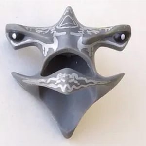 May include: A gray, stylized mask with a triangular top and two eye-like features. The mask has silver decorative patterns and a central opening. The design is symmetrical and appears to be made of a solid material.