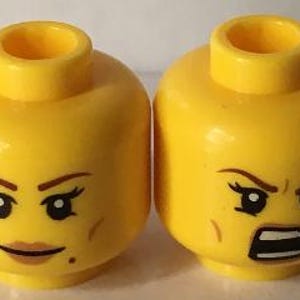 May include: Two yellow plastic toy figure heads with cylindrical neck studs. The left head features brown eyebrows, black eyelashes, and a neutral expression with brown lips. The right head displays furrowed eyebrows and an open, angry mouth.
