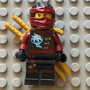 Może przedstawiać: Czerwono-czarna minifigurka Lego z czerwoną bandaną i złotym mieczem. Minifigurka ma czarny pasek i czerwoną szarfę. Minifigurka nosi czarną koszulę z białym i niebieskim wzorem.