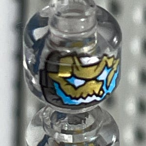 Peut inclure: Une tête de figurine Lego en plastique transparent avec un motif de crâne doré et bleu. Le crâne a un œil blanc et un motif bleu et blanc sur la mâchoire inférieure.