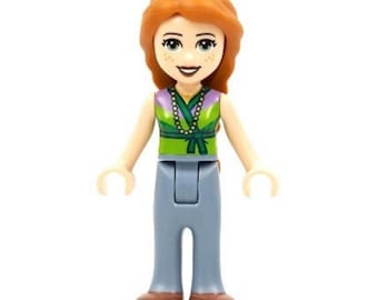 Puede incluir: Una figura de juguete de plástico con cabello naranja, una blusa sin mangas verde y morada con un collar estampado y pantalones azul claro. La figura tiene un tono de piel claro, pecas y una expresión sonriente con ojos y boca oscuros.