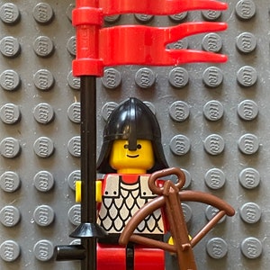 May include: A red and black Lego minifigure knight holding a crossbow. The knight is wearing a black helmet with a red plume, a chainmail shirt, and red pants. The knight is standing next to a black flagpole with a red flag.