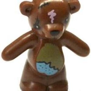 May include: A small brown teddy bear figurine with outstretched arms. The bear has a light purple marking on its forehead and a blue and green patch on its belly. It has black eyes and a stitched detail on its face.