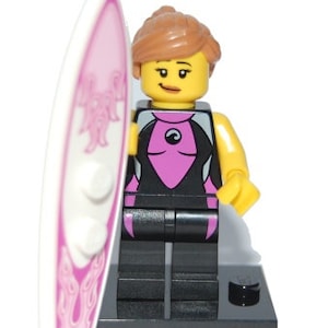 May include: A Lego minifigure of a female surfer holding a pink surfboard with white and pink designs. The figure is wearing a black wetsuit with a pink top and a black and white design on the chest. The figure has brown hair and a yellow face with a smile.
