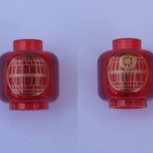 Pode incluir: Duas cabeças Lego vermelhas com um design dourado da Estrela da Morte. A Estrela da Morte é uma estação espacial fictícia da franquia Star Wars. As cabeças são feitas de plástico transparente com um acabamento brilhante. O design está centrado na frente da cabeça.