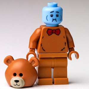 May include: A blue Lego minifigure with a sad face wearing a brown teddy bear costume with a red bow tie. The minifigure is holding a brown teddy bear head.