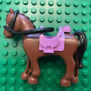 May include: A brown plastic toy horse with a black bridle and a purple saddle. The horse is standing on a green surface.
