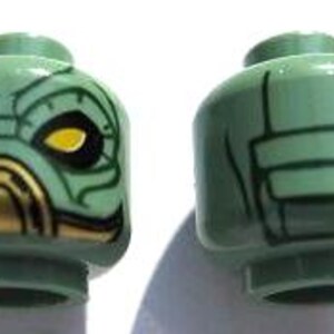 May include: Two green plastic toy heads. One head features a detailed face with yellow eyes and a gold-colored mouth. The other head has a design on the back. Both heads are for a toy figure.