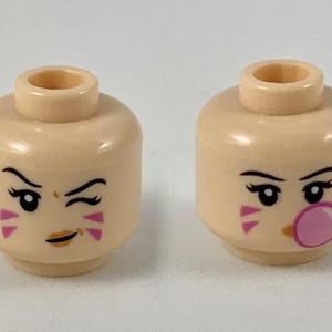 Op de afbeelding: Twee lichtgekleurde Lego minifiguren hoofden met verschillende uitdrukkingen. Het hoofd aan de linkerkant heeft een knipperend oog en een gouden mond. Het hoofd aan de rechterkant heeft een roze kauwgom in de mond en een verraste uitdrukking.