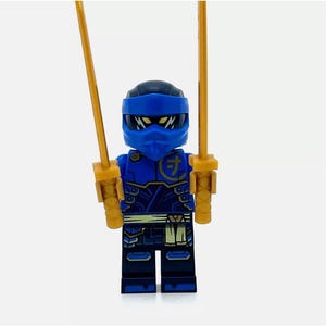 Może przedstawiać: Niebieska minifigurka Lego z czarną maską i złotymi mieczami. Minifigurka ma na sobie niebiesko-czarny strój z złotymi akcentami i złotym pasem. Minifigurka ma symbol na klatce piersiowej, który mówi "力".
