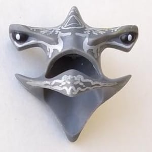 May include: A gray, stylized shark-shaped pendant with silver accents. The pendant features two eye-like details and decorative patterns. The design is symmetrical, with a pointed bottom and a central opening. The pendant is likely made of a durable material.