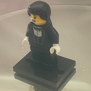 Può includere: Una minifigure Lego con un abito nero, camicia bianca e capelli neri. La figura ha un viso giallo e si trova su una piattaforma nera. Sorride leggermente.
