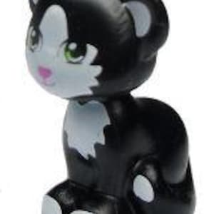 Puede incluir: Un juguete de gatito negro y blanco con ojos verdes y nariz rosa. El gatito está sentado con las patas metidas debajo del cuerpo.