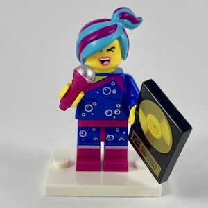 May include: A Lego minifigure of a singer with a blue and purple hairstyle, holding a pink microphone and a black record. The singer wears a blue jumpsuit with white bubble accents and pink accents. The record has a gold disc and the text "AWESOME".