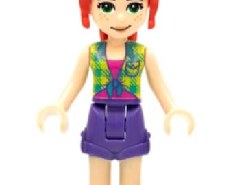 Puede incluir: Una minifigura de Lego Friends Emma. La figura tiene cabello rojo, ojos verdes y pecas. Viste un chaleco a cuadros, una camiseta rosa, pantalones cortos morados y zapatos azules. La figura está de pie con los brazos extendidos.