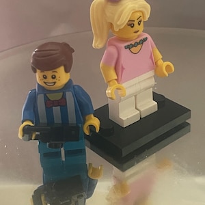 Puede incluir: Dos minifiguras de Lego. Una tiene cabello castaño, pecas y una camisa azul a rayas, sosteniendo un objeto negro. La otra tiene cabello rubio, una camisa rosa, pantalones blancos y un lazo morado. Están sobre una base negra.