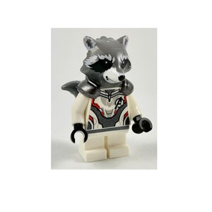 Puede incluir: Una minifigura Lego blanca de Rocket Raccoon de Guardianes de la Galaxie. La figura tiene una cabeza de mapache gris con ojos negros y una nariz roja. La figura lleva un traje blanco y gris con una pieza de pecho negra y roja.