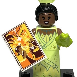 Puede incluir: Una minifigura LEGO verde y dorada de Tiana de la película de Disney La Princesa y la Rana. Lleva un vestido verde con detalles dorados y una corona. Sostiene una rana verde en su mano derecha y un boleto para Tiana's Place en su mano izquierda. La minifigura está de pie sobre una base LEGO negra con las palabras "Disney 100" impresas en ella.