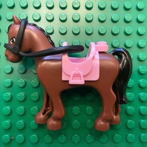 May include: A brown toy horse with a black mane and tail, wearing a black bridle and a pink saddle. The toy horse is made of plastic and is designed for play. The horse is standing on a green surface.