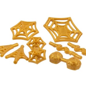 May include: A collection of gold-colored plastic toy pieces, including spiderwebs, lightning bolts, and other decorative shapes. The pieces are arranged on a white background, suggesting a Halloween or fantasy theme.