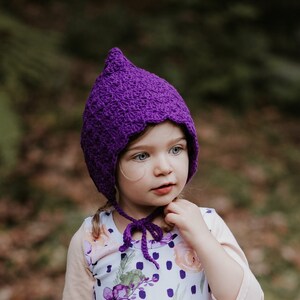 Puede incluir: Una niña pequeña con un gorro de crochet morado y un vestido blanco con estampado floral morado y lunares blancos.