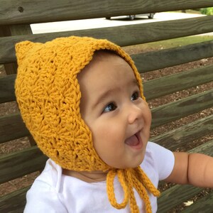 May include: A baby wearing a yellow crocheted bonnet with a tie. The bonnet has a pointed top and a scalloped edge.