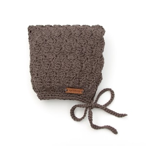 May include: A brown crocheted baby bonnet with a tie. The bonnet has a textured pattern and a small leather tag that says "Apricot & Co."