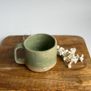 May include: A ceramic mug with a speckled sage green glaze on the upper portion and a natural beige clay base. The mug features a sturdy handle and a cylindrical shape, accompanied by a small cluster of white daisy-like flowers with yellow centres.