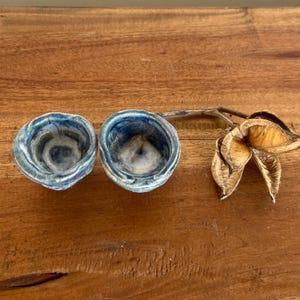 May include: Two small, handmade ceramic bowls with a textured, blue and white glaze. A dried, open seed pod with a tan and brown colour is next to the bowls. The items are on a wooden surface.