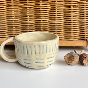 Op de afbeelding: Een witte keramische mok met een blauw-wit gestreept patroon. De mok heeft een afgeronde vorm en een handvat.