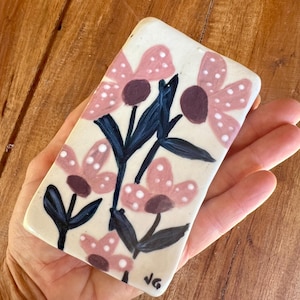 May include: Rectangular ceramic dish with a floral design. The flowers are pink with white polka dots and dark purple centres. Dark blue stems and leaves complete the design. The dish is off-white with a glossy finish. The initials "JG" are in the bottom right corner.