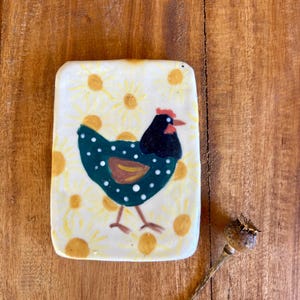 Pode incluir: Uma telha de cerâmica retangular com um desenho de galinha pintado à mão. A galinha é verde-azulado escuro com bolinhas brancas, uma cabeça preta e uma crista vermelha. O fundo é creme com detalhes de flores amarelas. Uma cápsula de semente seca está ao lado da telha.