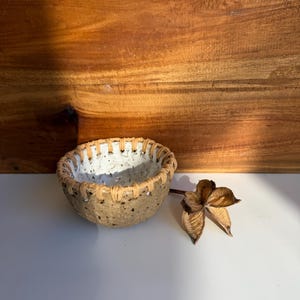 May include: A small, round, speckled ceramic bowl with a rough, tan-coloured exterior and a smooth, white interior. The rim of the bowl is wrapped in a light tan cord, creating a decorative stitched pattern around the top edge of the vessel.