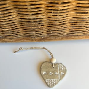 May include: A beige ceramic heart ornament with the word "mama" inscribed on it. The ornament is attached to a twine string with a wooden bead.
