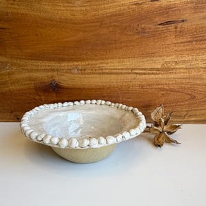 May include: A ceramic bowl with a beige exterior and a glossy white interior. The rim of the bowl is decorated with a continuous row of small, rounded white clay beads. A dried, star-shaped botanical seed pod rests beside the bowl on the surface.