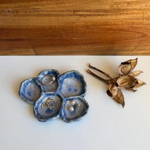 May include: A blue and white ceramic trinket dish with five rounded compartments, featuring a glossy finish and irregular edges. The dish contains a silver ring, a pair of silver hoop earrings, and a pair of pearl stud earrings.