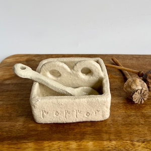 May include: A rectangular, off-white, textured ceramic pepper cellar with a matching spoon resting inside. The front of the container features the word pepper embossed in lowercase letters. Two circular openings are visible on the back wall.
