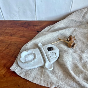 May include: White ceramic salt and pepper dish with a spoon. The dish is shaped like a flower with three sections. One section has salt, another has peppercorns, and the third is empty. The dish is sitting on a beige linen cloth.