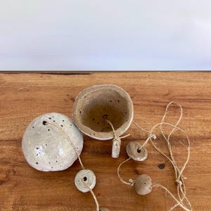 Pode incluir: Um conjunto de vasos suspensos de cerâmica feitos à mão. O conjunto inclui uma esfera redonda, salpicada, esbranquiçada, uma tigela pequena e várias contas pequenas e redondas enfiadas em barbante natural. O vaso é projetado para pendurar.