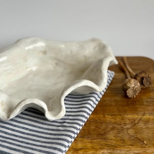 May include: A white ceramic bowl with an irregular, wavy, and organic rim shape. The bowl has a glossy, off-white finish with subtle texture. It is placed on a folded white and navy blue striped cloth napkin, which rests on a wooden surface.