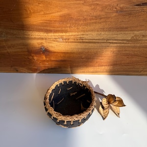 May include: A small, round, black bowl with a textured surface, featuring a decorative rim and exterior stitching made from light tan natural fibre cord. A single dried, brown, star-shaped botanical element rests beside the bowl.