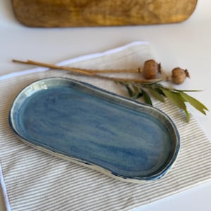 Puede incluir: Una bandeja ovalada de cerámica con un esmalte azul brillante y sutiles variaciones de tono. La bandeja tiene un borde ligeramente elevado e irregular y una base poco profunda. El borde exterior muestra una base de cerámica de color blanco roto.