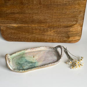 Pode incluir: Uma bandeja oval de cerâmica com duas pequenas alças, apresentando uma superfície esmaltada com tons misturados de verde sálvia, rosa suave e creme. As bordas têm um acabamento texturizado e irregular, acompanhada por flores brancas secas.