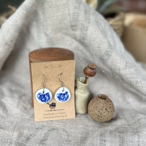 May include: A pair of white ceramic earrings with a blue and white design. The earrings are handmade in Victoria, Australia. The earrings are hanging from a card with the text "Jess Grey Art" and "handmade in Victoria" and a website address.
