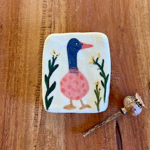 May include: A square ceramic tile with a hand-painted duck design. The duck has a blue head, pink body with polka dots, and orange beak and feet. Green plants and yellow flowers surround the duck. A dried seed pod rests on a wooden surface.