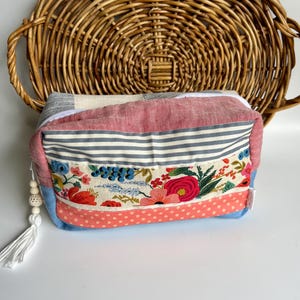 Puede incluir: Una bolsa de cosméticos rectangular de tela patchwork con cierre de cremallera. La bolsa está hecha de diferentes telas, incluyendo rosa, azul, blanco y estampado floral. La bolsa tiene un borla blanca y una cuenta blanca.