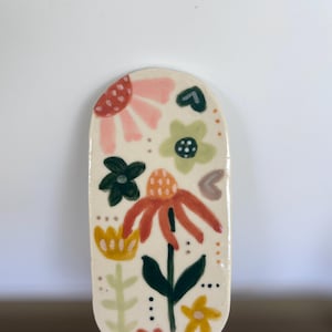 May include: An oval ceramic trinket dish with a cream-coloured base featuring hand-painted floral motifs in shades of pink, orange, yellow, and green. The design includes various stylised flowers, leaves, and small dots, with the initials JG at the bottom.